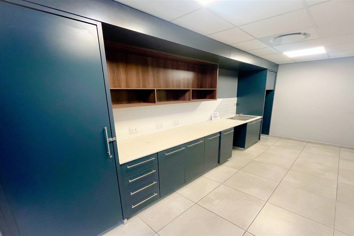 A modern office kitchenette featuring dark blue cabinetry, light countertops, integrated shelving, and a stainless steel sink with tiled flooring.