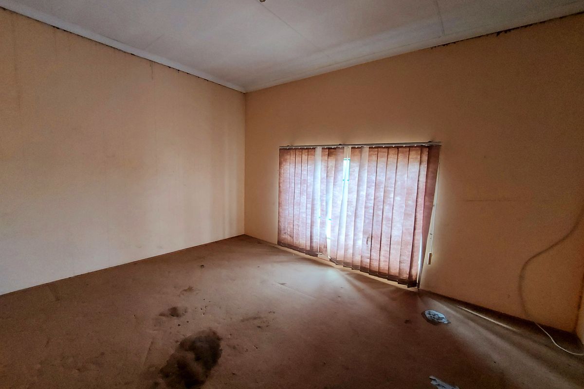 An empty bedroom featuring worn carpeting, plain walls, and vertical window blinds, requiring cosmetic updates and refurbishment.
