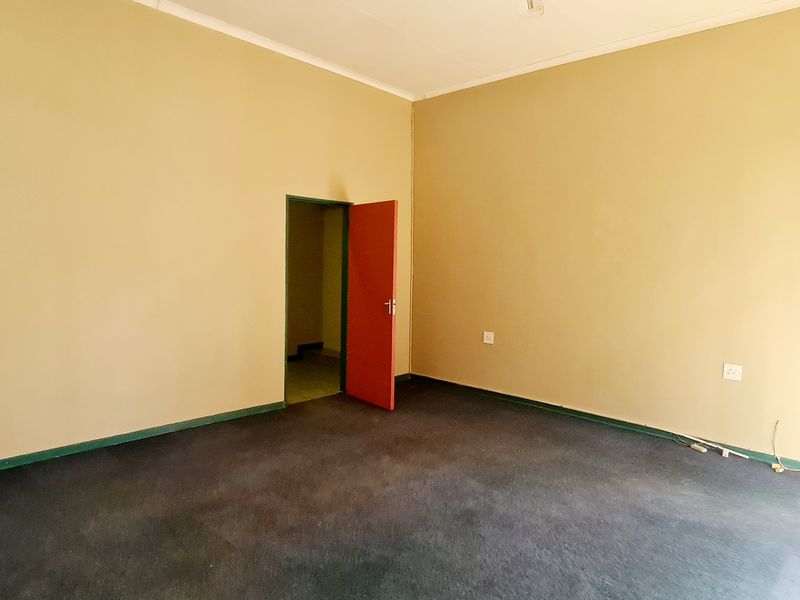 An empty office space featuring carpeted flooring, neutral wall tones, and access to an adjacent room via a red door.