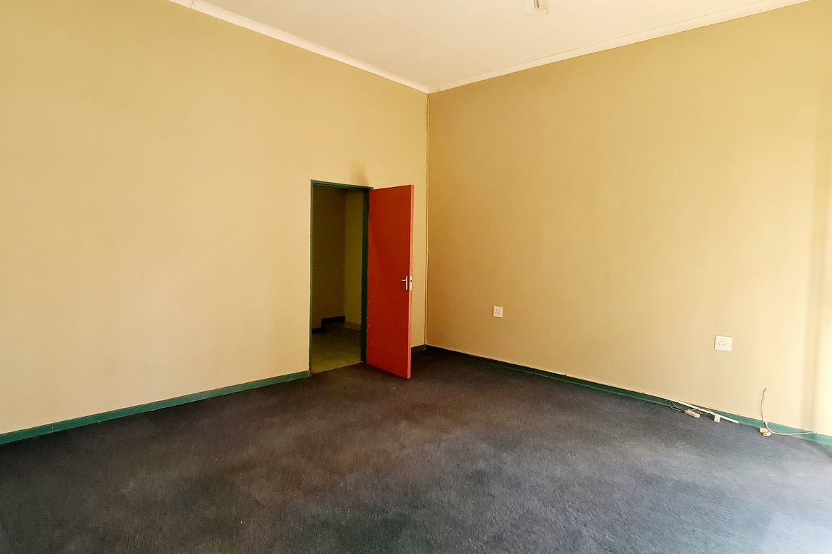 An empty office space featuring carpeted flooring, neutral wall tones, and access to an adjacent room via a red door.