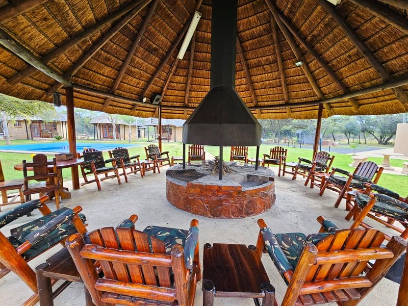 A spacious outdoor thatched lapa featuring a central stone fireplace with a large metal extractor hood, perfect for social gatherings and entertainment.