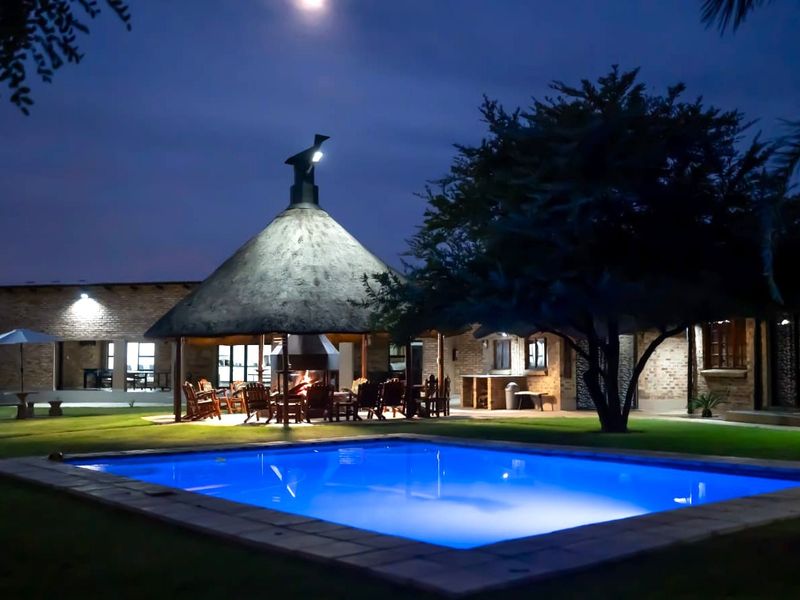 Night view of a well-maintained farm property featuring a swimming pool, a large thatched lapa with a built-in braai, and an outdoor entertainment area.