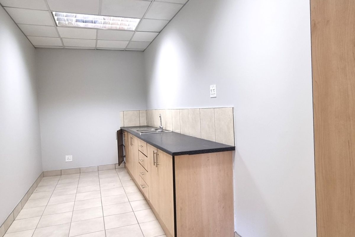 A functional office kitchenette featuring light wood cabinetry, a dark countertop, a stainless steel sink, and tiled flooring with a drop ceiling.