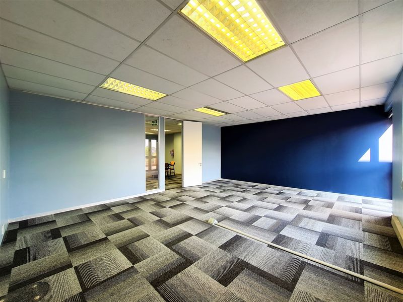 A spacious, empty office area featuring modern carpet tiles, a suspended ceiling with recessed lighting, and a glass partition door.