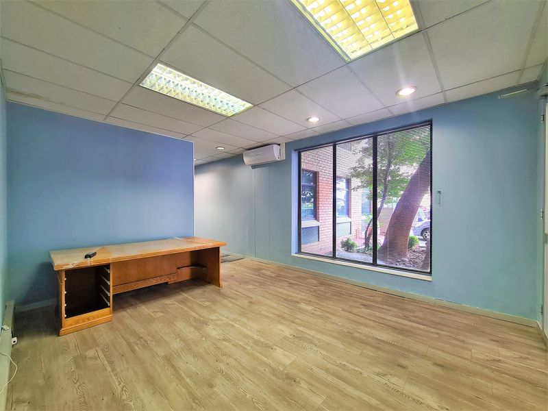 A bright, empty office space featuring laminate flooring, a suspended ceiling with recessed lighting, and a split-system air conditioner.