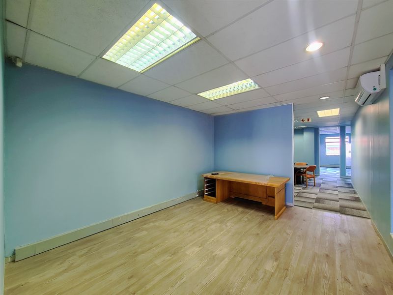 A spacious, empty office area featuring laminate flooring, a suspended ceiling with recessed lighting, and a wall-mounted air conditioning unit.