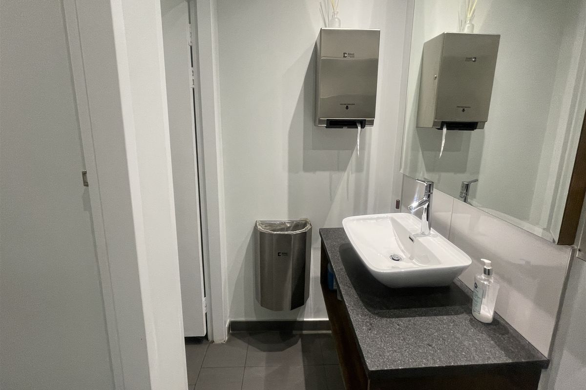 A clean office restroom featuring a modern vessel sink, stone countertop, wall-mounted paper towel dispenser, and stainless steel waste bin.