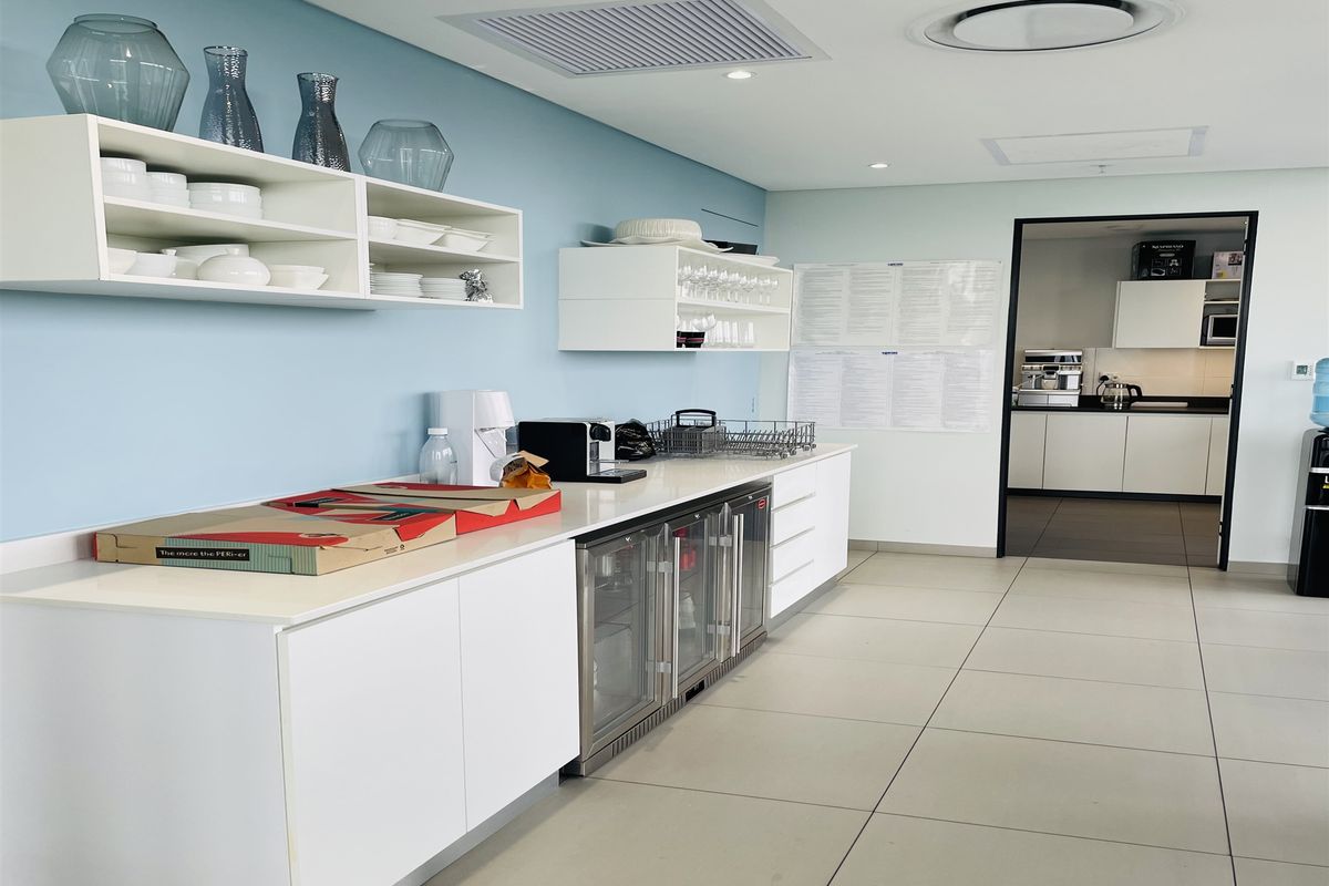A modern office kitchenette featuring white cabinetry, integrated under-counter beverage refrigeration, and open shelving, with a secondary prep area visible through the doorway.