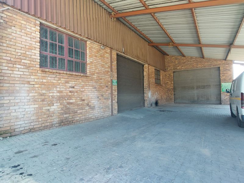 Industrial exterior rear view featuring brick facade, corrugated metal roofing, and multiple roller shutter doors with paved access area.