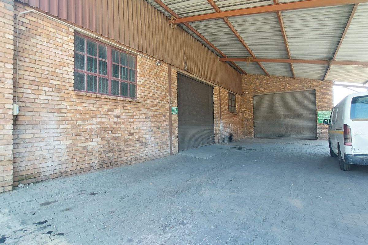 Industrial exterior rear view featuring brick facade, corrugated metal roofing, and multiple roller shutter doors with paved access area.