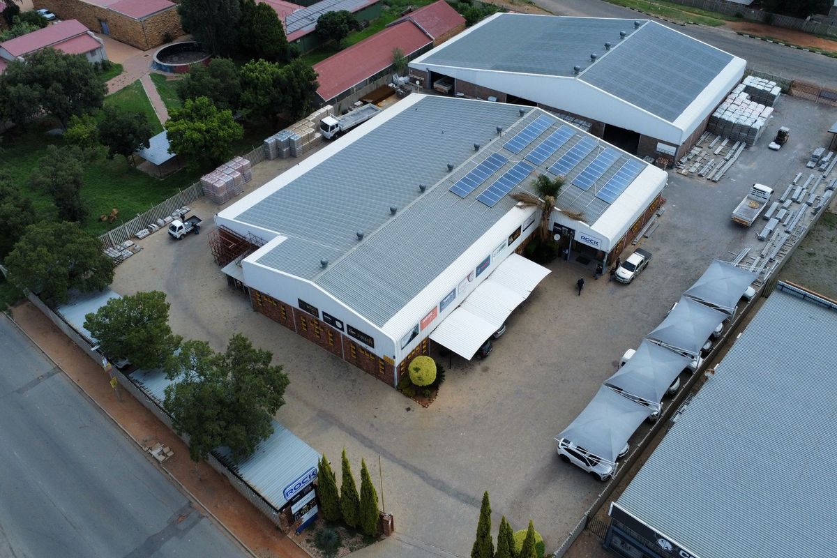 Aerial view of an industrial property featuring a large warehouse with solar panels, paved parking area, and shade cloth carports.
