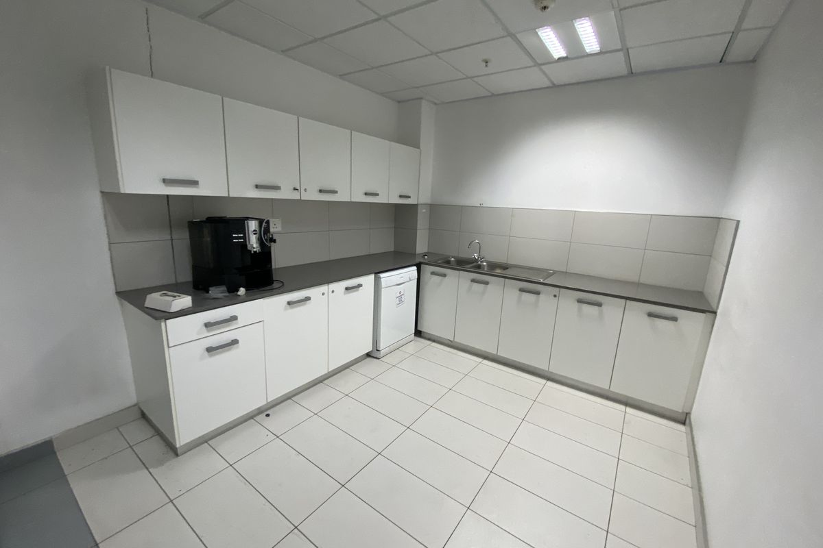 A functional office kitchenette featuring white cabinetry, a stainless steel sink, tiled flooring, and a drop ceiling with integrated lighting.