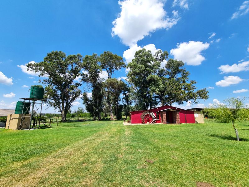 A wide view of the farm property featuring a red building, large mature trees, and elevated water storage tanks on a steel stand.