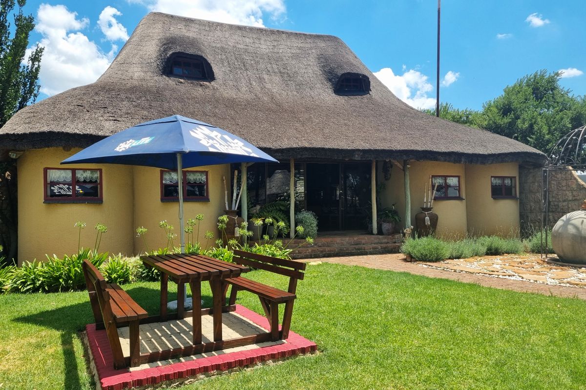 A charming thatched-roof farmhouse featuring a covered patio area, well-maintained garden, and paved outdoor seating space.