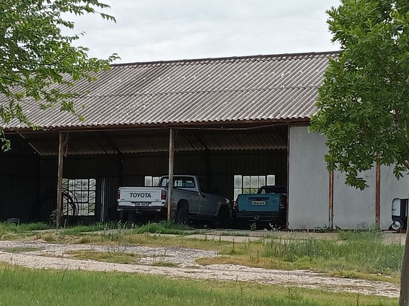 An open-sided farm shed or garage structure featuring a corrugated metal roof and steel support columns, suitable for vehicle storage.