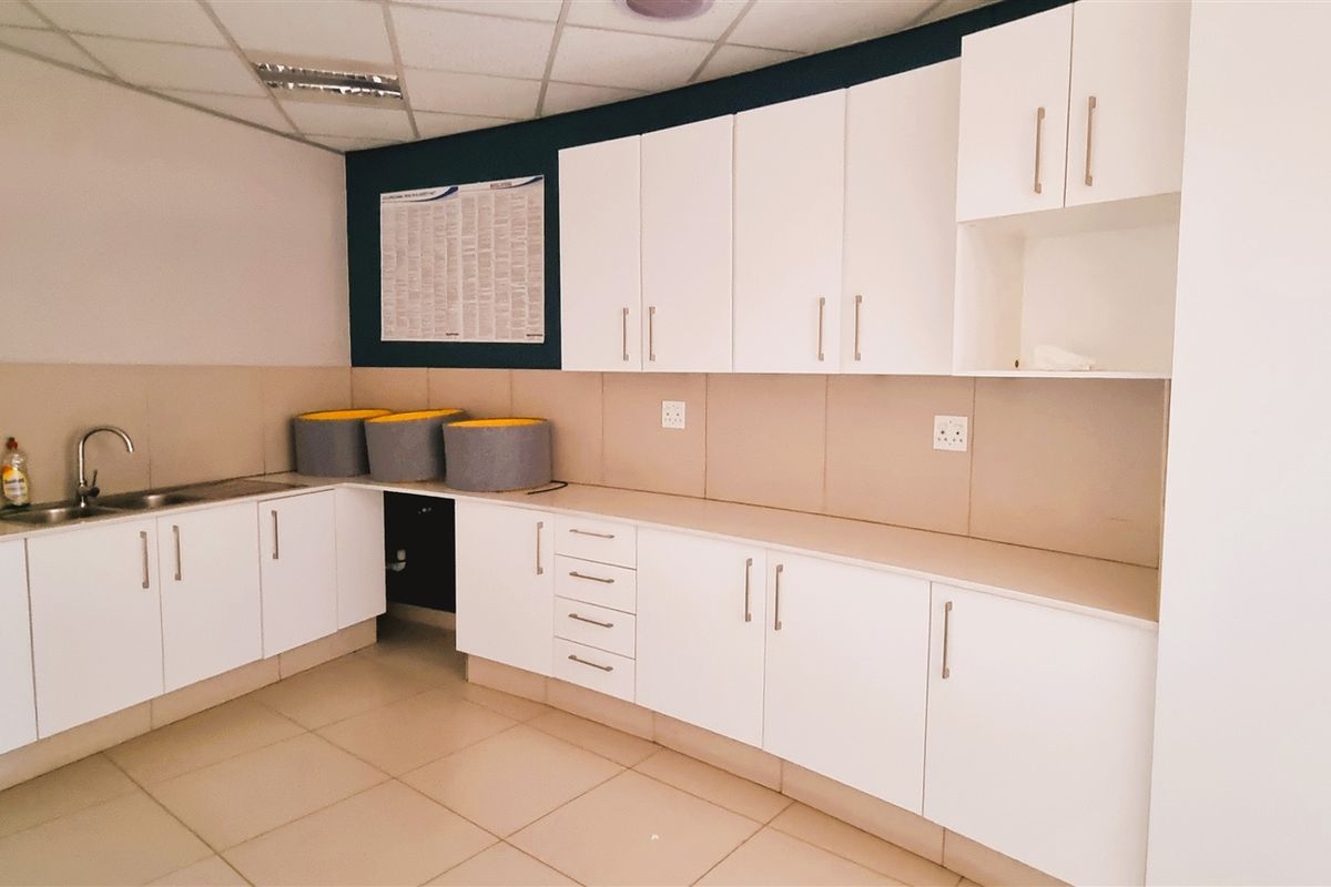 A functional office kitchenette featuring white cabinetry, tiled flooring, a stainless steel sink, and ample counter space for staff use.