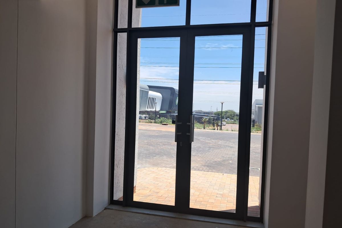 A bright, modern entrance area featuring double glass doors leading to a paved exterior space, complete with an overhead emergency exit sign.