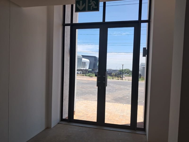 A bright, modern entrance area featuring double glass doors leading to a paved exterior space, complete with an overhead emergency exit sign.