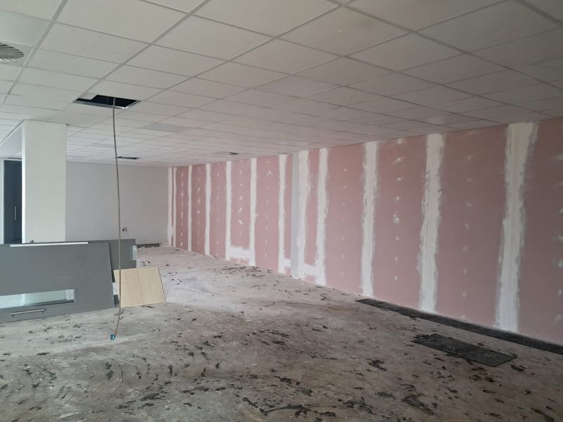 An unfinished office space featuring exposed drywall, a suspended ceiling with missing tiles, and bare flooring, currently undergoing renovation.