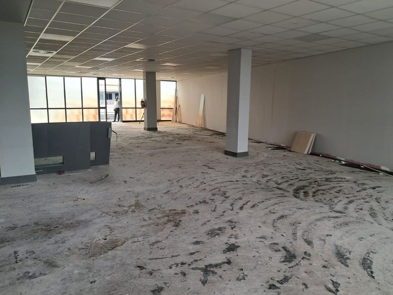 An open-plan office space featuring a suspended ceiling, structural columns, and large windows, currently requiring flooring installation and interior finishing.