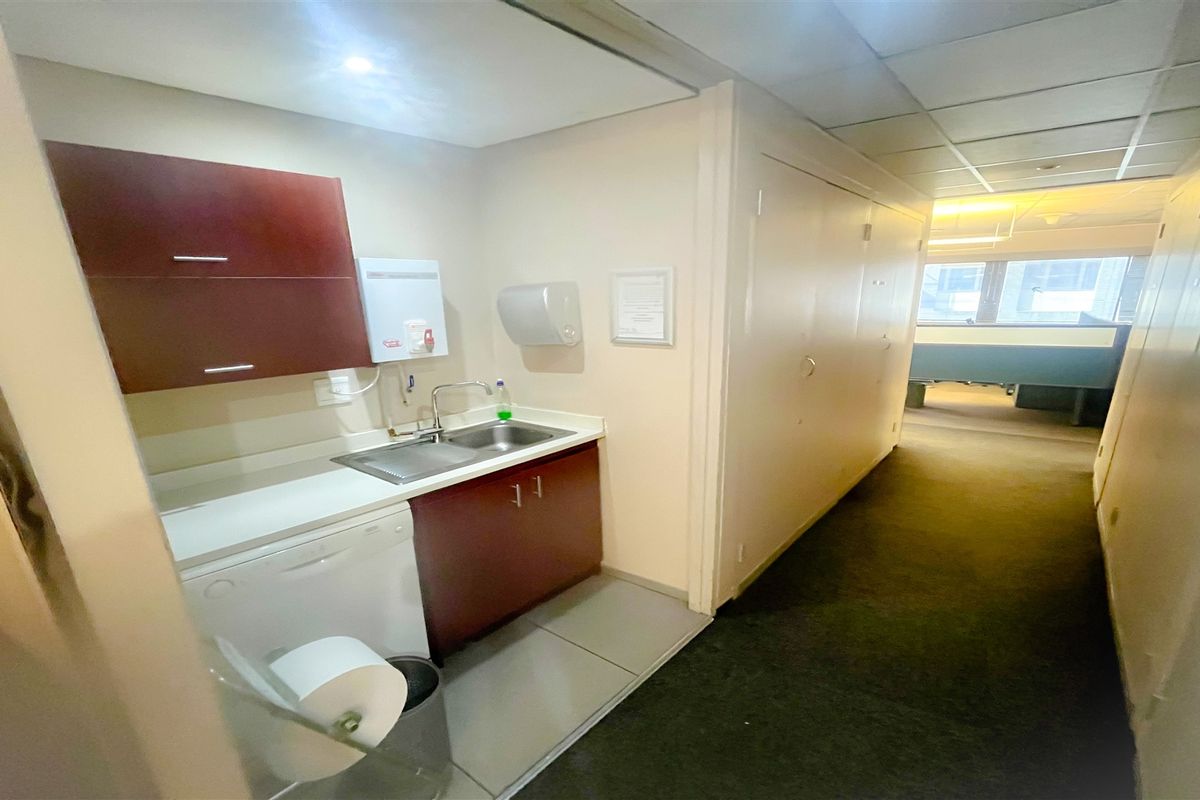 A compact office kitchenette featuring built-in cabinetry, a stainless steel sink, and tiled flooring, situated adjacent to a carpeted office hallway.