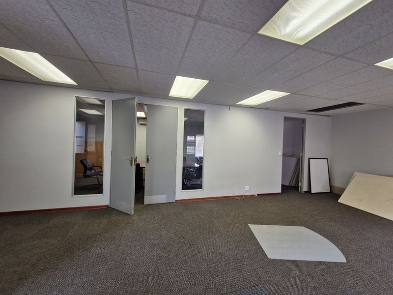 An open-plan office space featuring a suspended ceiling with recessed lighting, carpeted flooring, and glass partition panels providing visibility into adjacent rooms.