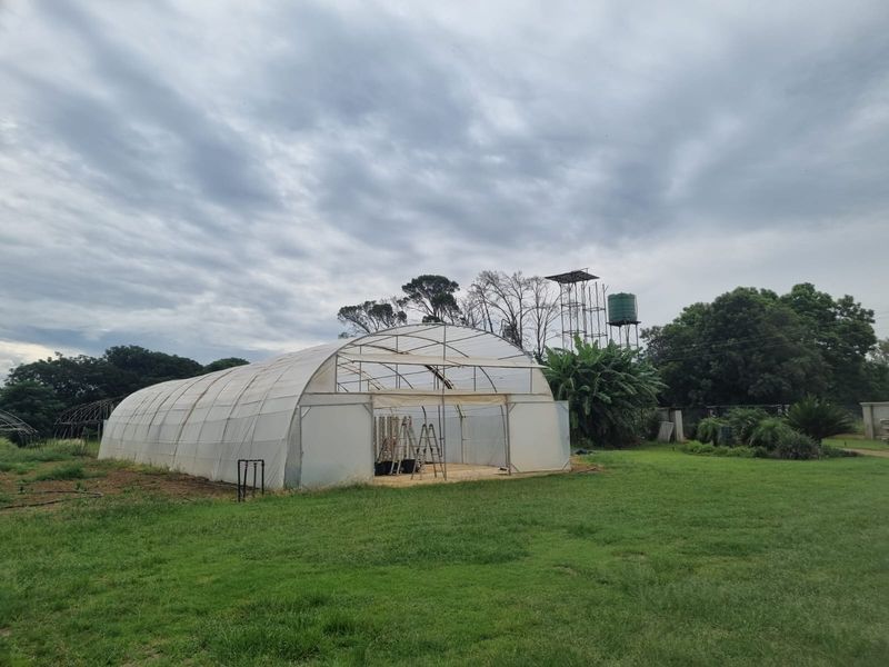 A large agricultural greenhouse structure situated on a grassy plot, accompanied by an elevated water tank and tower infrastructure.