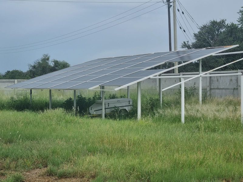 A ground-mounted solar panel array installation situated within a fenced farm area, featuring a metal support frame and concrete perimeter walling.