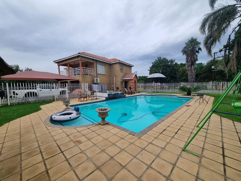 A spacious outdoor pool area featuring a large swimming pool, paved decking, and a two-story brick farmhouse with a balcony.