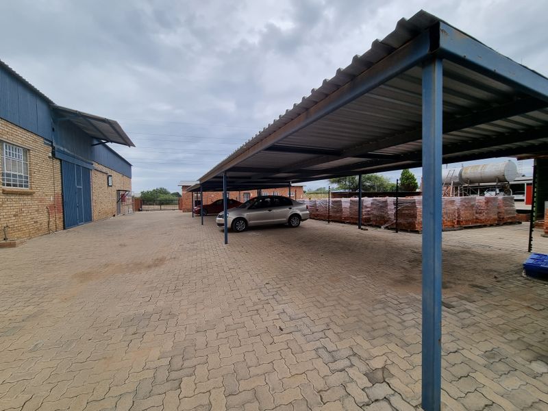 Commercial exterior area featuring a large paved yard, covered carport structure, and brick building facade.