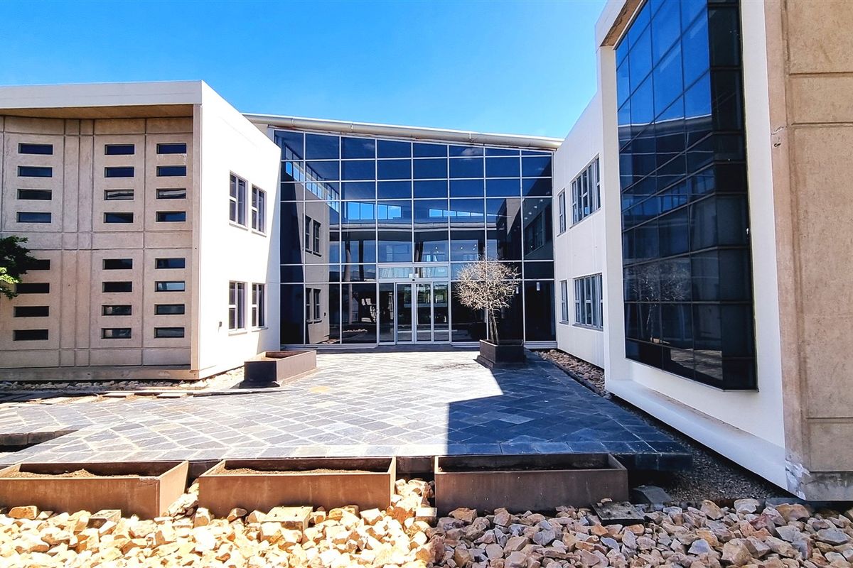 Modern office building exterior featuring a large glass curtain wall facade, paved courtyard area, and structured landscaping.