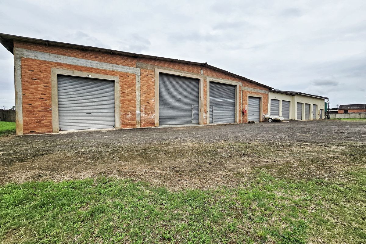 Exterior view of an industrial warehouse facility featuring multiple roller shutter doors and exposed brick construction with a gravel yard area.