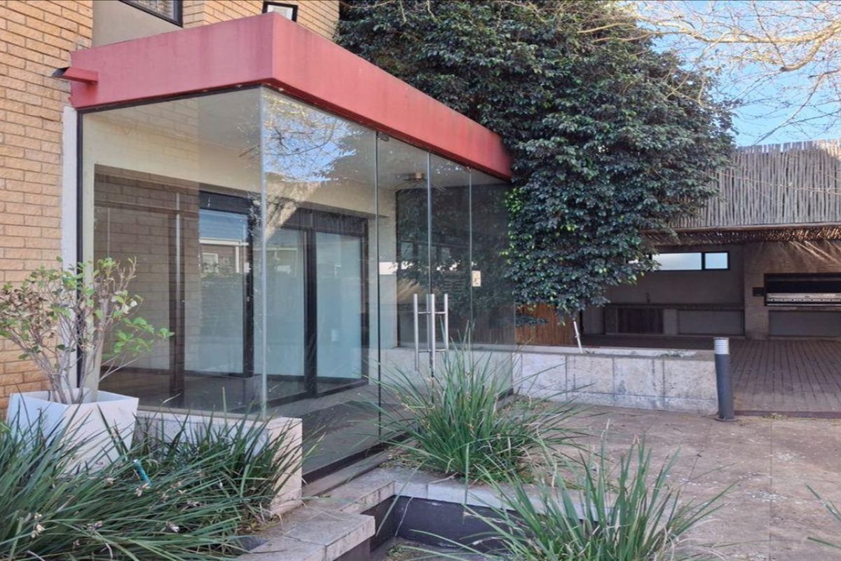 Rear exterior view featuring a glass-enclosed entrance, paved patio area, and a built-in braai under a thatched roof structure.