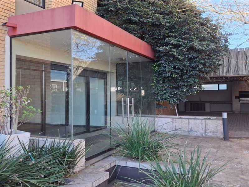 Rear exterior view featuring a glass-enclosed entrance, paved patio area, and a built-in braai under a thatched roof structure.