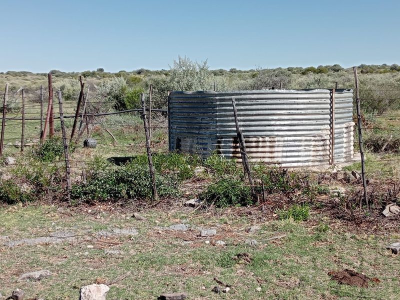 A weathered corrugated iron water tank situated in a rural farm setting, enclosed by aging wire fencing and metal posts.
