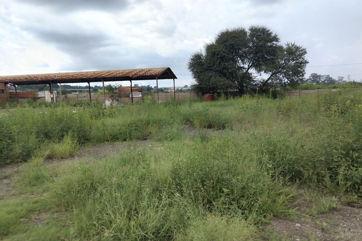 An industrial outdoor yard area featuring a large, rusted metal roof structure and perimeter walling, currently overgrown with vegetation.