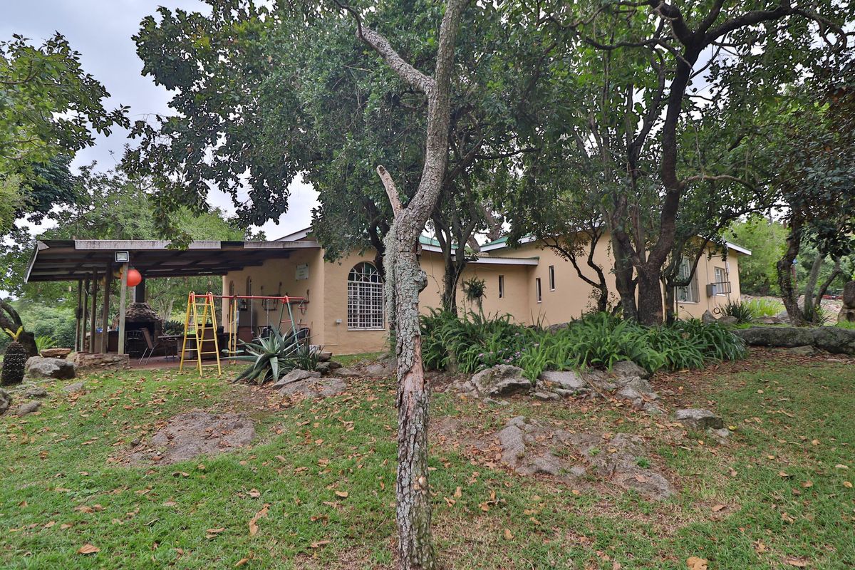 Rear view of the farm property featuring a covered patio with a built-in braai, set within a mature garden with established trees and rock features.