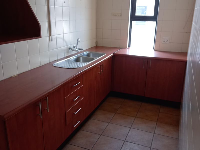 A functional office kitchenette featuring laminate countertops, built-in wooden cabinetry, a stainless steel double sink, and tiled flooring.