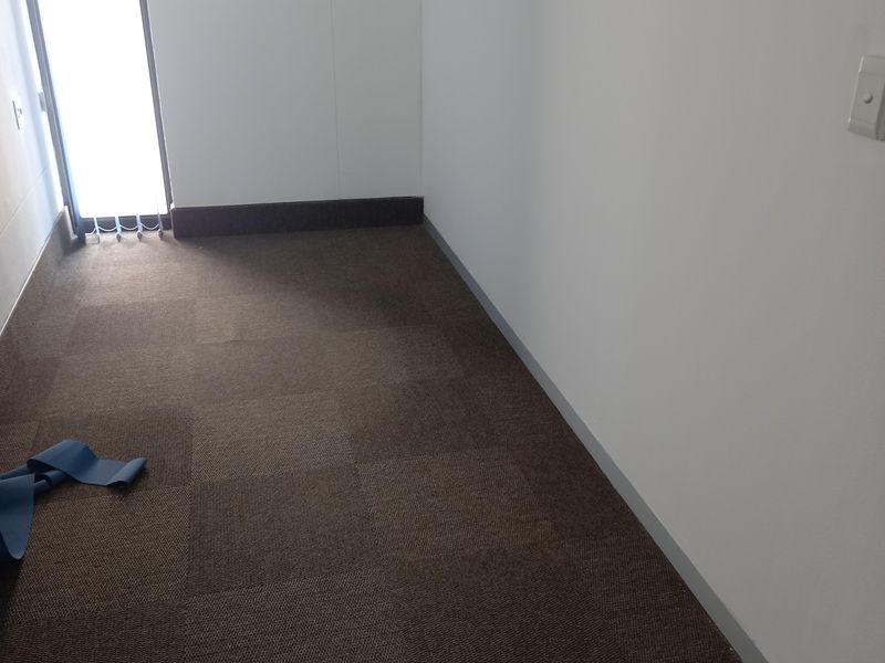 A vacant office space featuring neutral carpet tiles, white walls, and a tall window with vertical blinds.