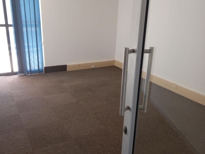A vacant office space featuring carpet tiles, a wall-mounted air conditioning unit, and a glass entrance door with vertical blinds.