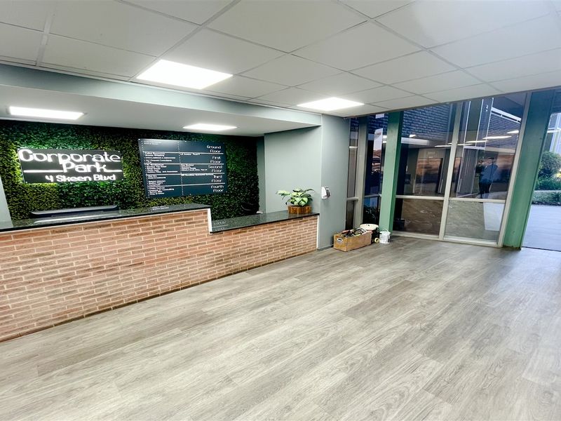 Modern office building entrance lobby featuring a brick-fronted reception desk, artificial green wall branding, and large windows providing natural light.