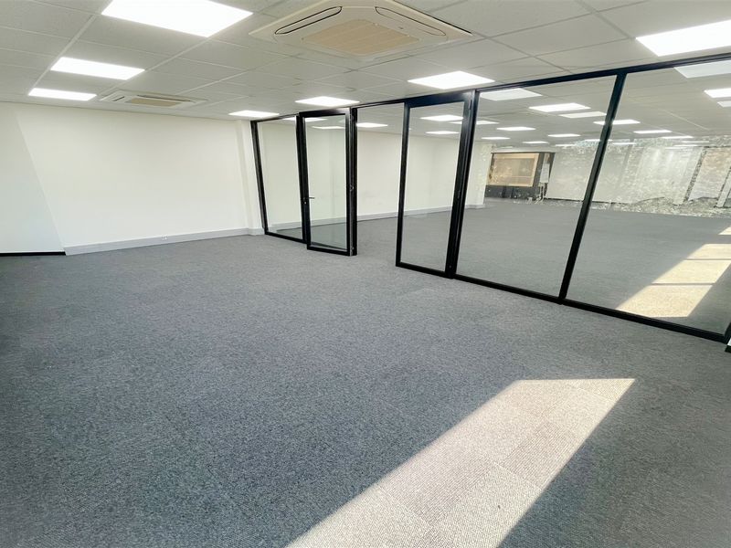 A modern, vacant office space featuring glass partition walls, carpeted flooring, and a suspended ceiling with integrated recessed lighting and air conditioning.