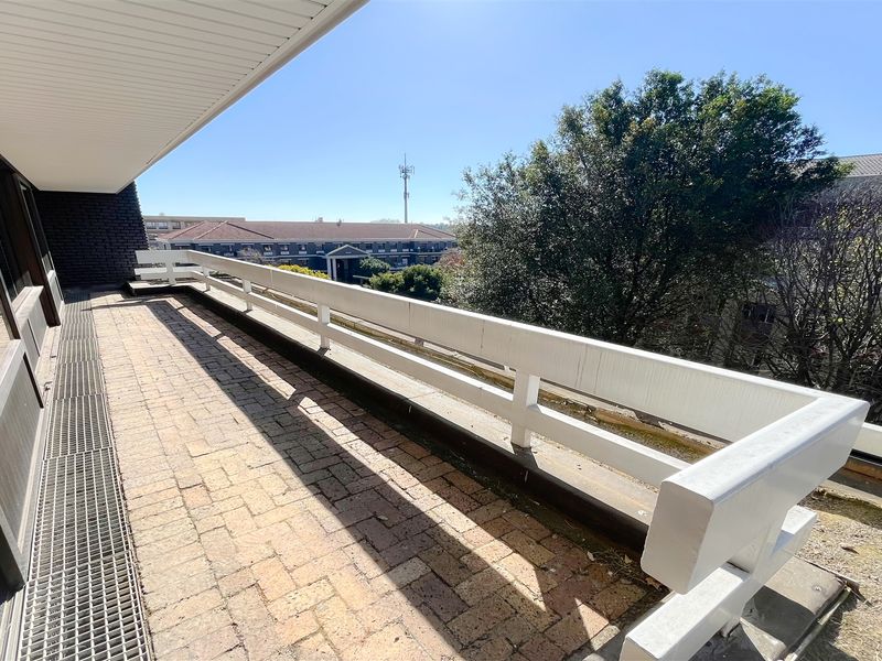 A spacious outdoor balcony featuring brick paving, a white metal balustrade, and a drainage grate, offering an elevated view of the surrounding office complex.