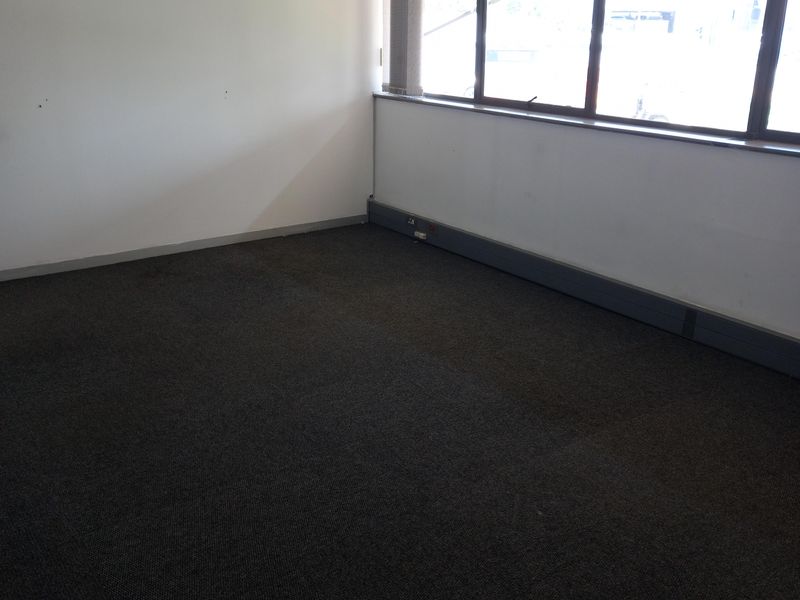 An empty office space featuring dark carpeting, a suspended ceiling, wall-mounted air conditioning, and large windows providing natural light.