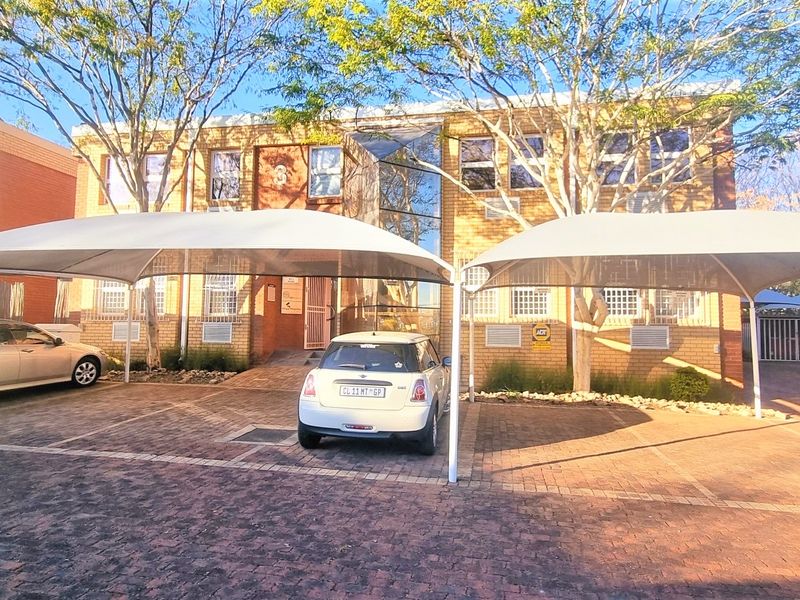 Front exterior view of a multi-story office building featuring a paved parking area with shade cloth carports and established trees.