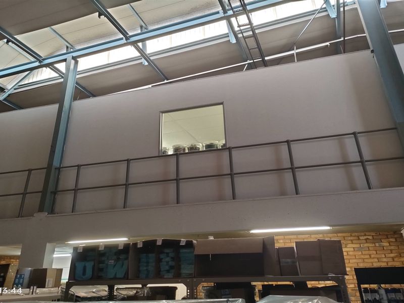 Industrial warehouse interior featuring a mezzanine level with metal railing, exposed steel structural beams, and natural light from overhead skylights.