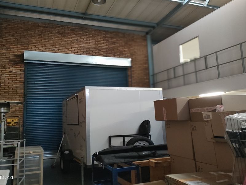 Industrial warehouse space featuring high ceilings with exposed steel beams, a large roller shutter door, and an elevated mezzanine level with metal railings.