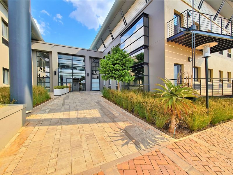 Modern office building entrance featuring a paved walkway, glass facade, and well-maintained landscaped garden beds.