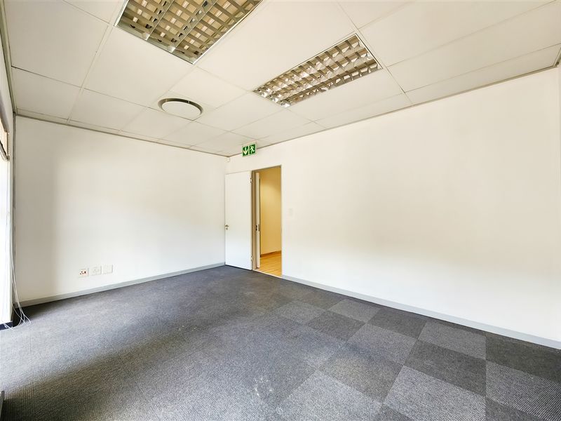 A clean, vacant office space featuring carpet tiles, a suspended ceiling with recessed lighting, and an emergency exit sign.