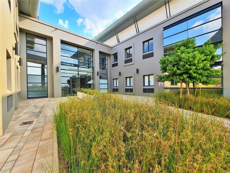 Modern office building exterior featuring a landscaped courtyard, paved walkways, and extensive glass facade elements.